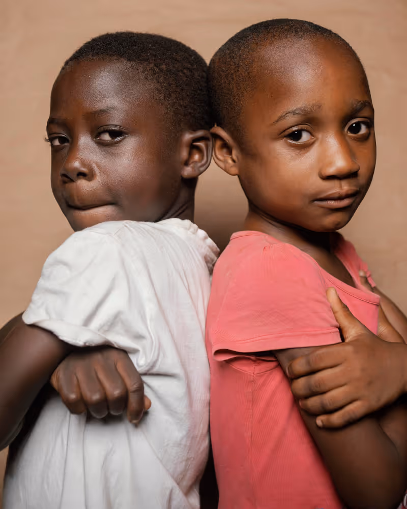 Two young children standing back to back with arms crossed, looking at the camera against a plain background.