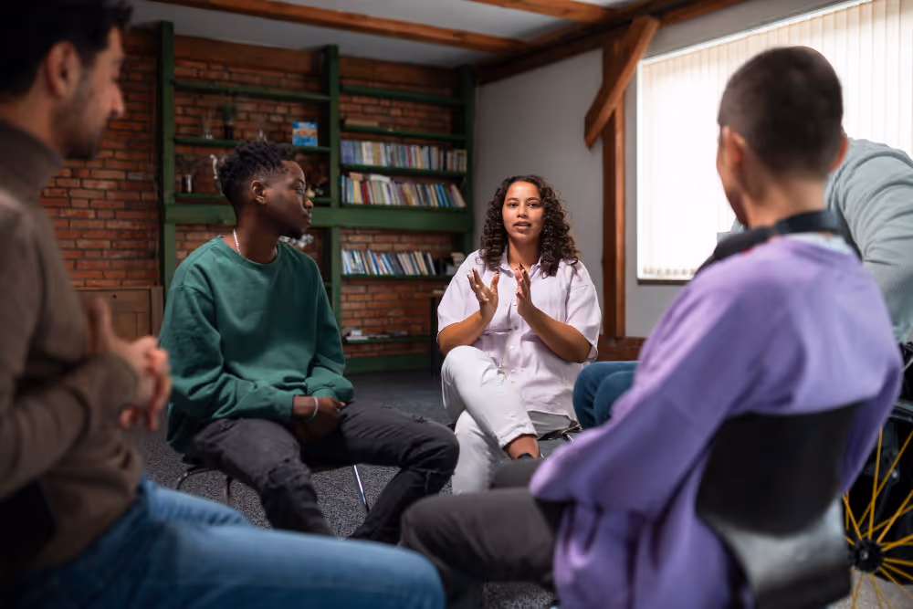 A group of diverse young adults sitting in a circle, engaged in a discussion in a cozy room with bookshelves and brick walls.