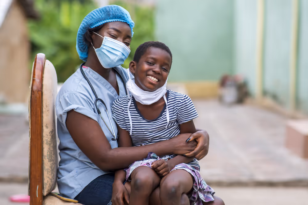 Smiling healthcare worker wearing a surgical mask and cap embraces a happy child with a face mask pulled down.