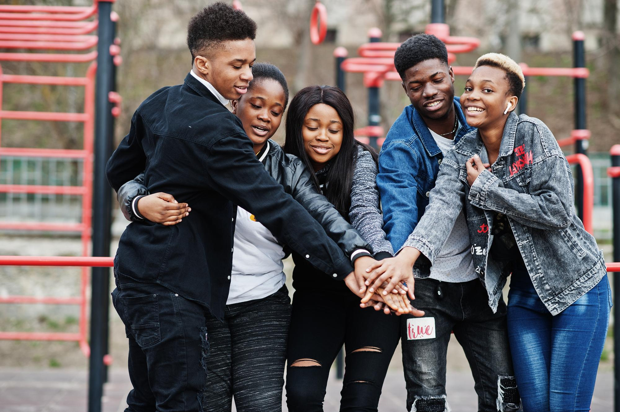 Five young people standing close together outdoors with their hands stacked in the center, smiling and showing friendship.