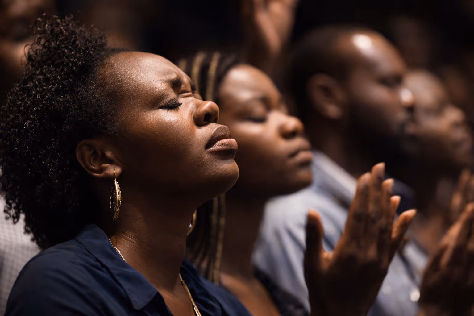 Close-up of a woman with closed eyes and raised hands in prayer or meditation, with others in a similar posture blurred in the background.