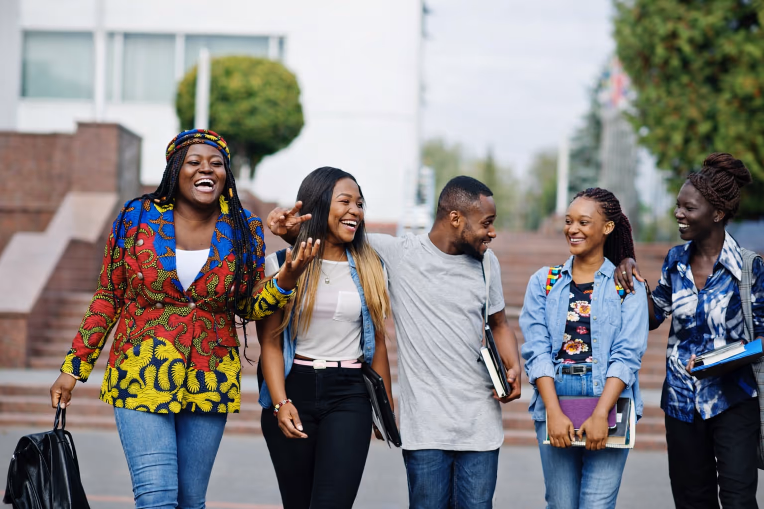 Group of five young adults smiling and walking together outdoors, carrying books and backpacks.
