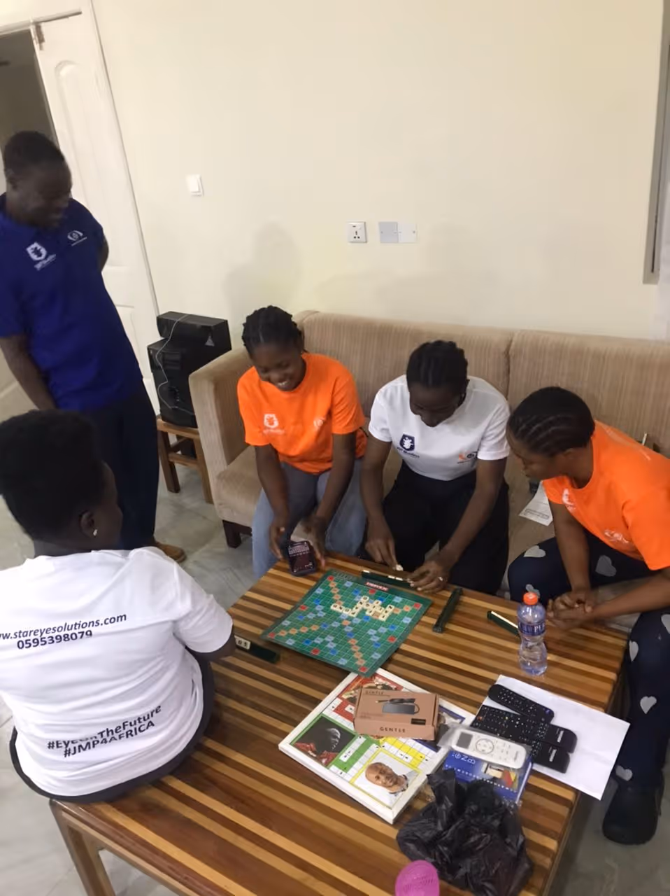 Four young women sitting around a striped wooden table playing Scrabble, one standing nearby, with various items including remotes and a water bottle on the table.