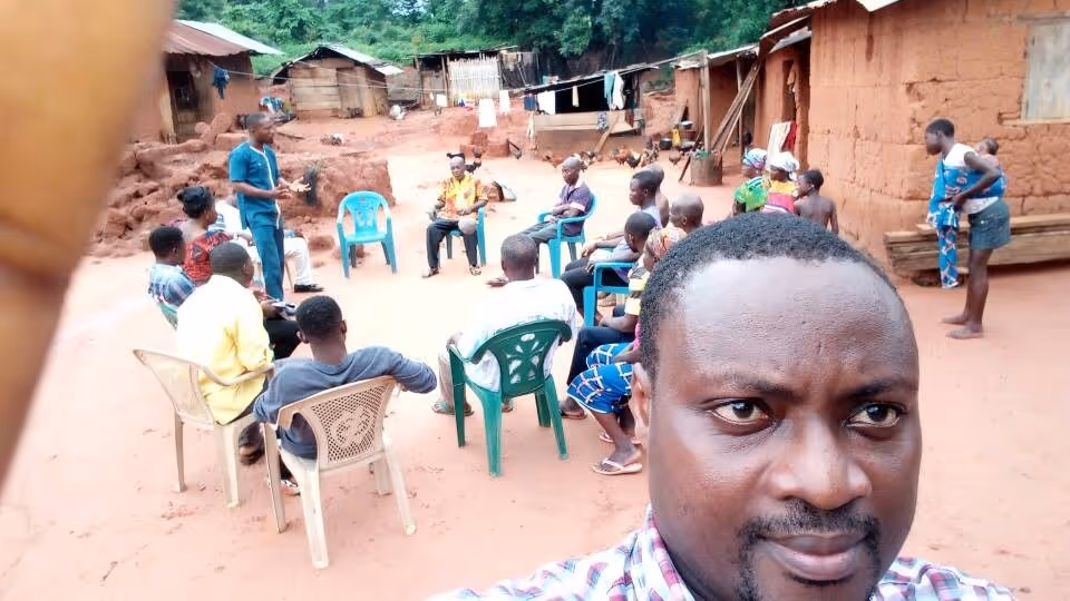 Group of people seated in plastic chairs arranged in a circle outdoors in a rural setting with mud houses, one man taking a selfie in the foreground.